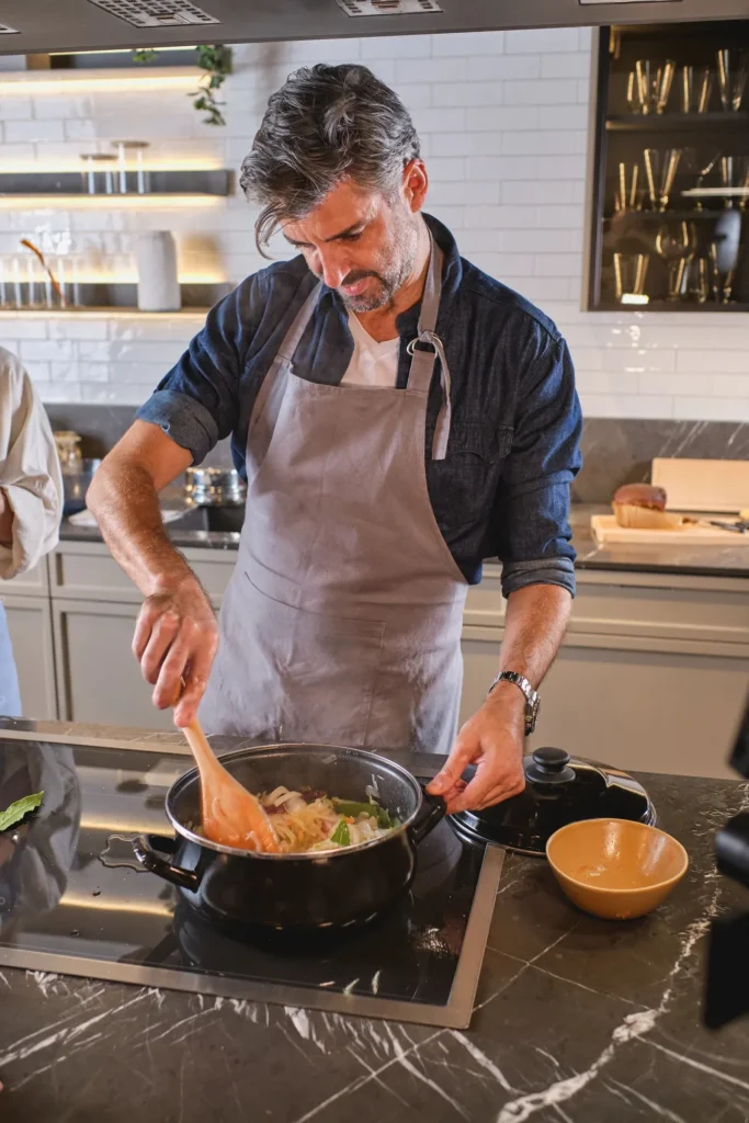 About me 1 Man in apron adding chopped meat to a pot while cooking in a modern kitchen